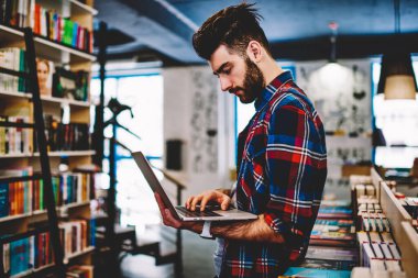 Düşünceli sakallı hippi adam dizüstü bilgisayar tutuyor ve modern kütüphanede kablosuz internet bağlantısı kullanarak cihaza uygulama yüklüyor. Netbook 'a yazılım yükleyen konsantre genç adam.