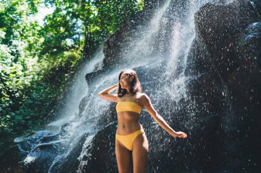 Güneşli bir yaz gününde, yeşil bitkilerin yanında durup bakışlarını kaçırırken mayo giyen genç bir bayan.