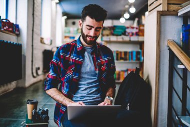 Neşeli hipster blogcu günlük kıyafetlerle gülerken kendi web sitesinden kablosuz internet bağlantısı kullanarak dizüstü bilgisayarda oturarak yayın yapıyor.