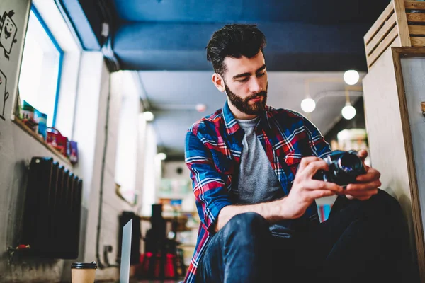 Professional bearded photographer dressed in casual wear holding vintage camera and making setting spending time in studio.Creative young man programming on device sitting in coworking space