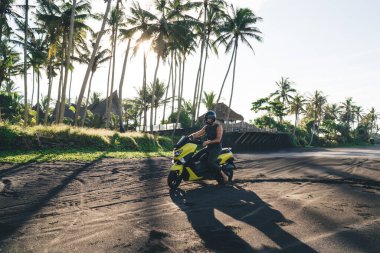 Side view full length male biker in summer wear and yellow helmet riding motorcycle on black sandy path along tropical palm in sunny day