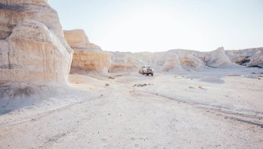 Güneşli bir günde, açık mavi gökyüzüyle arka planda giden kamyonla düz kumtaşı oluşumları olan kuru çöl manzarasının resmedilebilir görüntüsü.
