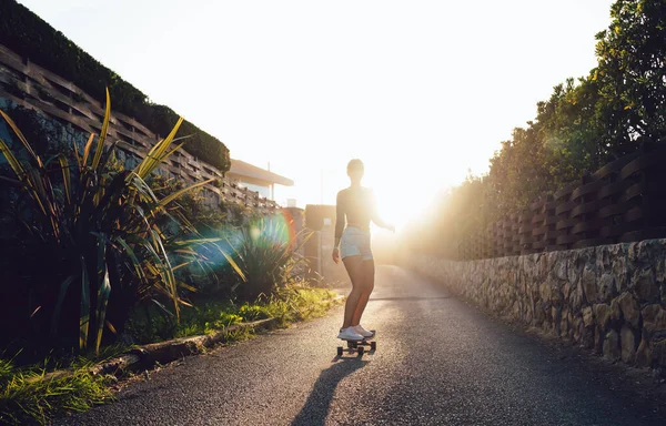 Full body of anonymous athletic female skater in trendy outfit ride longboard on asphalt path against cloudless sky at sunset