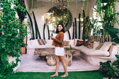 Yazlık elbisesi içinde gülümseyen Asyalı kadın yeşil çimlerin üzerinde duruyor ve kameraya bakarken cep telefonuna bakıyor.