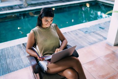 Yukardan, günlük giyinen kız öğrencilerin elinde dizüstü bilgisayarla oturup yüzme havuzunun yanında otururken klavyeye yazı yazması.