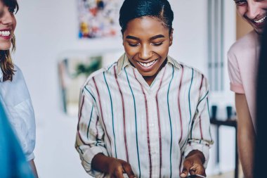 Gülümseyen, coşkulu Afrikalı Amerikalı kadın elinde kağıtlarla elinde arkadaşlarıyla gülüyor ve ışık tasarım odasında birlikte kalıyorlar.