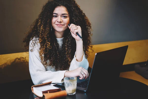 Kıvırcık saçlı yetenekli bir öğrenci kafeteryada serbest çalışırken kameraya bakıyor.