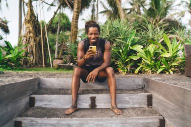 Tüm vücudum, günlük kıyafetlerle gülümseyen siyah bir erkek tropikal bahçede taş basamaklarda oturuyor ve internette sörf yaparken kameraya bakıyor.