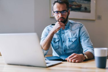 Gömlek içinde konsantre olmuş bir erkek kameraya bakıyor çenesinden el ele tutuşuyor laptopla masada oturuyor ve bir fincan kahve içiyor.