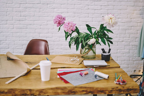 Takeaway coffee and vase of flowers placed on wooden table near sewing supplies left by seamstress after work on new clothes