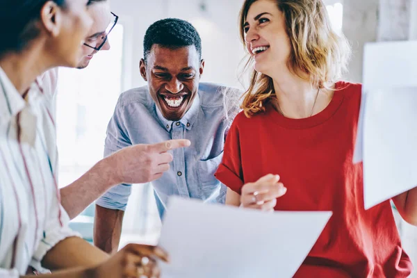 Smiling cheerful laughing intelligent young multiracial coworkers in casual apparel checking documents standing at table in modern light loft office space