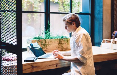 Gündüz vakti rahat iç mekanı ve büyük penceresi olan bir kafede çalışırken laptop kullanan düşünceli bir öğrencinin yan görünüşü