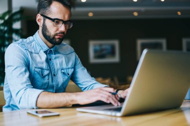 Güncel giysiler içinde konsantre sakallı bir adam akıllı telefonuyla masada oturmuş çağdaş iş yerinde çalışırken ekran tarayıcı bilgisayarına bakıyor.