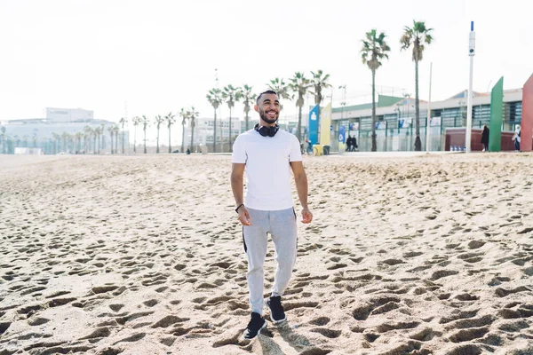 Positive bearded ethnic man in sportswear with headphones resting on sandy beach and walk on seashore after training at daytime