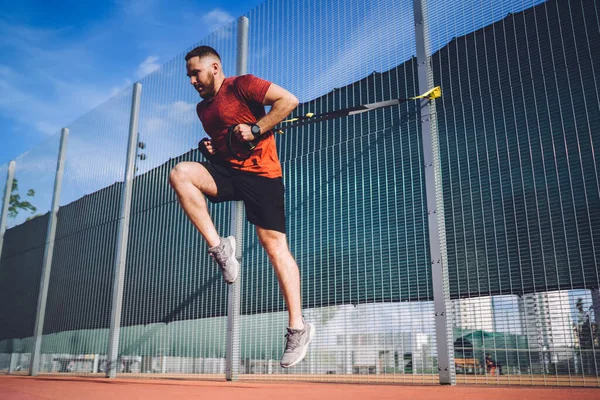 From below of full body sportsman jumping during intense exercises and looking away while practicing suspension training on sports ground