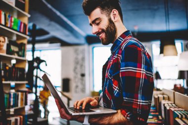 Elinde dizüstü bilgisayar tutan sakallı neşeli genç adam ve modern kütüphanede kablosuz internet bağlantısı kullanarak uygulama yükleyerek. Netbook online sohbet Pozitif hipster adam