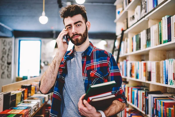Good looking hipster guy looking at camera while making telephone call at college campus, casual dressed male student choosing books for preparing to exam and talking with friend via smartphone