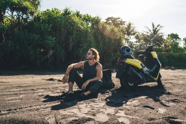 若い自信に満ちた男性のヒップスターのフルボディ離れて見て 熱帯リゾートでの旅行中に一人でリラックスしながらバイクの近くの砂の上に座って — ストック写真