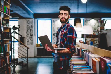 Modern dizüstü bilgisayarı tutan ve ücretsiz internet kullanarak uygulama yükleyen şık yenilikçi öğrenci. Modern kütüphanede güncel profilini güncelleyen sakallı genç adam.