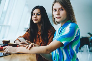 Genç bayan arkadaşlar, kafeteryada gündüz vakti kahve ve akıllı telefonlar eşliğinde günlük kıyafetlerini giyerken kameraya bakarlar.