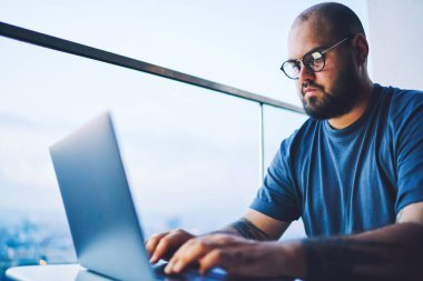 Düşük açılı, rahat tişörtlü, hafif çalışma alanında çalışırken açık dizüstü bilgisayarıyla masada oturan sakallı bir erkek işçi.