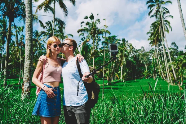 陽気若いです男と女でカジュアル服とサングラス撮影で写真とともに自撮り棒接吻しながら緑のトロピカルガーデンを歩く — ストック写真