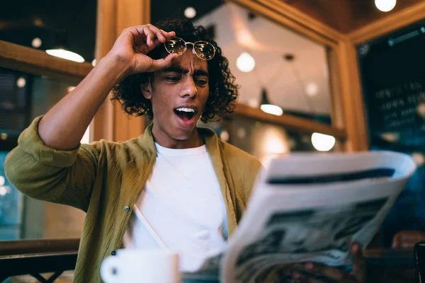 Emotional curly hipster guy amazed with latest news from printed media taking off eyewear while reading