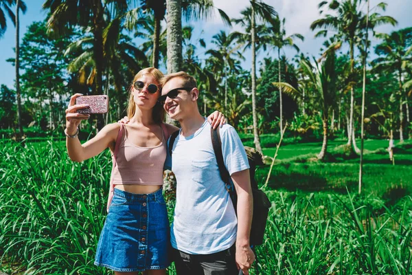 Güneş gözlüğü takmış, sırt çantalı, gülümseyen ve tropik bahçede cep telefonundan selfie çeken mutlu bir genç adam ve kadın.