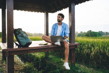 Şık giyinmiş düşünceli genç erkek yazar doğulu çardakta dizüstü bilgisayarla oturarak yeni bir makale yazarken pitoresk kırsalda ilham alıyor.