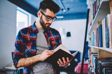 Sıradan giyinmiş sakallı genç adam elinde edebiyat kitabı tutuyor ve kütüphanede duran sayfalar hakkında faydalı bilgiler arıyor.