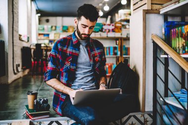 Gündelik giysiler içinde, modern kütüphanedeki kablosuz internet bağlantısını kullanarak dizüstü bilgisayarda ders çalışması yapan dalgın sakallı genç adam. İnternet sitelerinde bilgi arayan öğrenci.