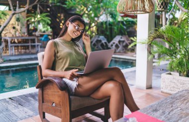 Gözlüklerini ayarlayan, bacak bacak üstüne atmış bir şekilde bilgisayarda yüzme havuzunun yanında bulanık arka planda çalışan zevkli bir kadın.