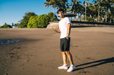 Aktif giyimli, kafası karışmış genç bir erkek atlet boş deniz kıyısında yürürken mesajlaşmak için cep telefonu kullanıyor ve kameraya bakıyor.