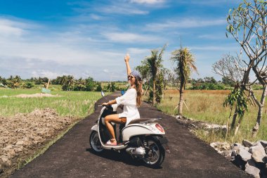 Tam vücut pozitifliği olan yaz kıyafetleri ve güneş gözlüklü gezgin motosiklet üzerinde oturuyor ve tropikal bir ülkede tatil yaparken poz veriyor.