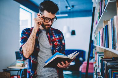Görme düzeltme için gözlük pensive akıllı hipster öğrenci elinde ders kitabı tutan ve ders yazmak için bilgi okuma. Kütüphanede ayakta rahat giyim konsantre genç adam