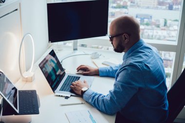 Bilgisayar ve tabletle masada çalışan ve modern ofiste dizüstü bilgisayar kullanan resmi kıyafet ve gözlüklü genç işadamının yan görüntüsü