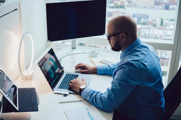 Bilgisayar ve tabletle masada çalışan ve modern ofiste dizüstü bilgisayar kullanan resmi kıyafet ve gözlüklü genç işadamının yan görüntüsü