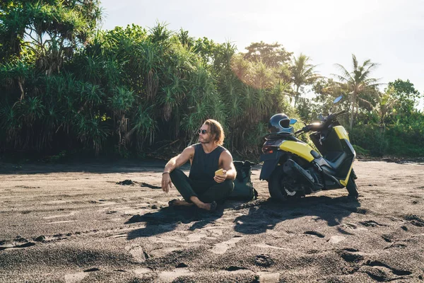 Full body of brutal male hipster with mobile phone in hand chilling on sand near scooter and looking away while messaging during trip