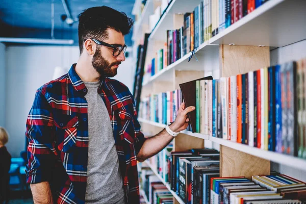戴眼镜的多愁善感的学生站在大书架上 为自己的课程选择文学 聪明的年轻人戴着眼镜 手里拿着一本书 准备在教育图书馆参加考试 — 图库照片