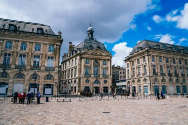 Bordeaux 'da bulutlu bir sonbahar gününde ortaçağ döneminden kalma tarihi binaların etrafında turistlerin dolaştığı eski asfalt bir meydan.