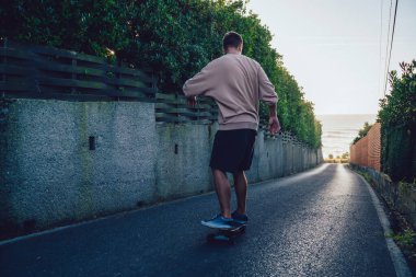 Spor kıyafetli, yeşil ağaçlara karşı boş yolda tek başına at süren ve antrenman yapan isimsiz genç bir erkek kaykaycının arka görüntüsü.