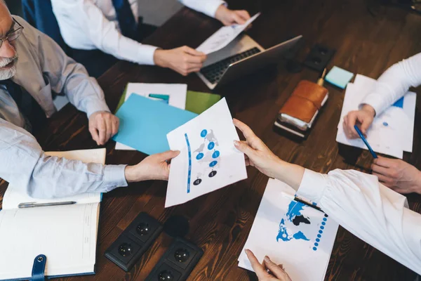 From above of group of crop anonymous intelligent business colleagues discussing information and diagrams on papers at table with laptop