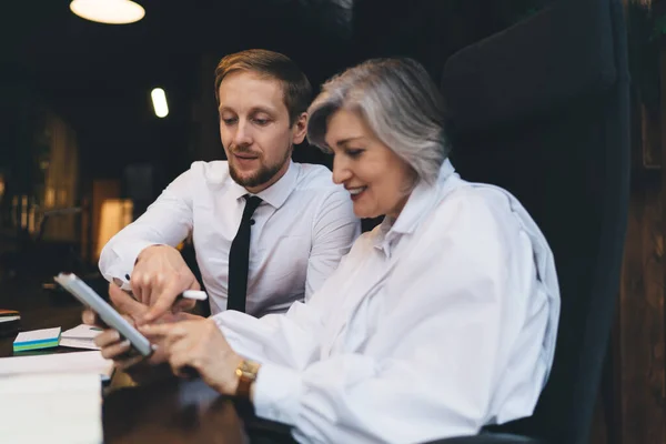 Satisfied business partners in formal outfits surfing internet on mobile phone while making plan for new business project in light workspace