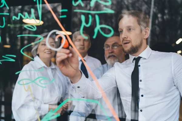 Group of businesspeople in formal outfits solving strategy while young bearded colleague writing on glass of window in conference hall