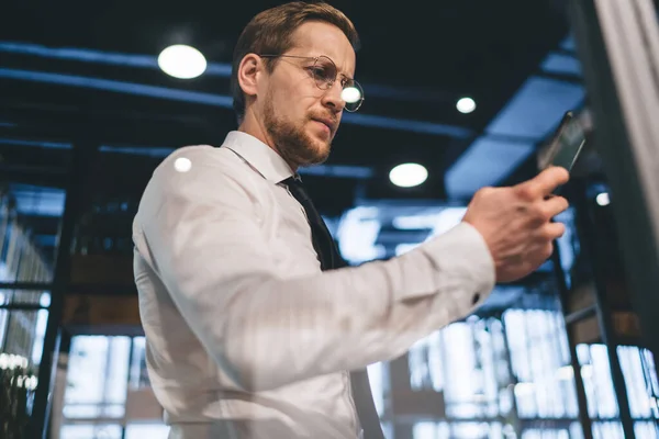 From below through glass of serious male in white shirt and tie checking notifications on mobile while standing in modern office with burning lamps