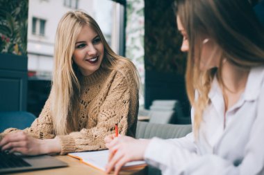 Kafedeki masada oturan gülümseyen genç bir bayan iş projesinde çalışırken iş arkadaşıyla konuşuyor ve dizüstü bilgisayar kullanıyor.