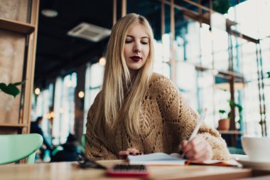 Düşük açılı, serbest giyinmiş, kırmızı rujlu bir kadın, ahşap masada oturuyor ve kafedeki deftere not alıyor.