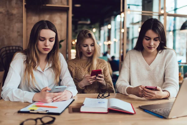 Focused female students in casual wear using smartphones while sitting at table with clipboard notebook and laptop in modern coffee shop