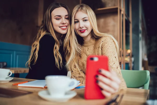 穿着休闲装笑着的女性朋友们坐在餐桌边的咖啡店里 一边在智能手机上自作主张 一边彼此寒战 — 图库照片