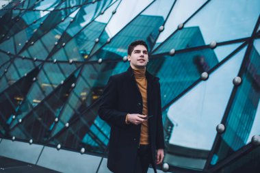 Akıllı telefonlu, kendinden emin genç bir çalışan haftasonunu dışarıda geçirirken diğer tarafa bakıyor ve modern binanın yanında duruyor.